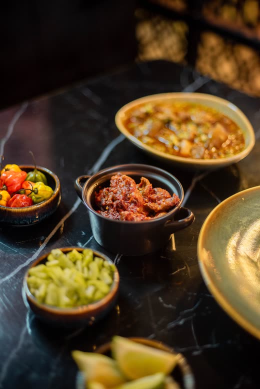 A spread on black marble — a bowl of stew with okra, small dishes of coloured peppers, chopped greens and lime wedges