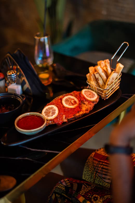 A long slate plate with a whole spiced fish, lemon slices, a wire basket of chips and two dipping sauces
