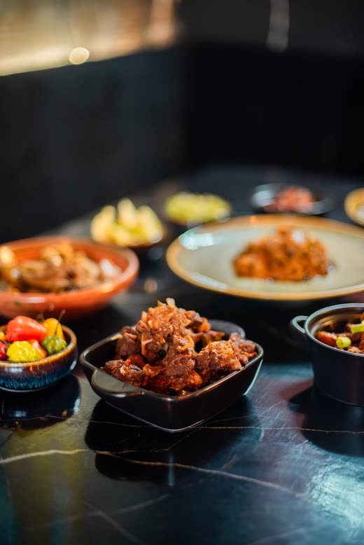 A square black dish of meat stew on a dark table, peppers and greens alongside