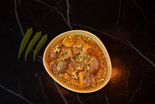 A single bowl of stew with meat and okra on black marble, three okra pods laid beside it