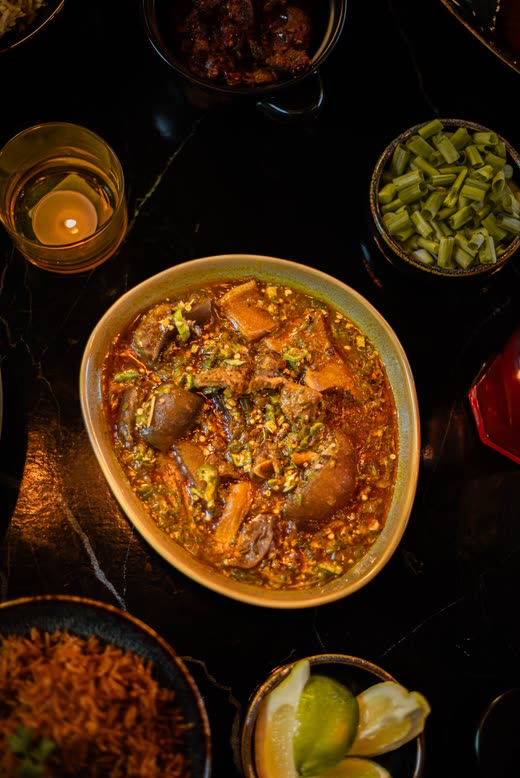 Seen from above — a wide bowl of stew with meat and greens, candles and side bowls around it