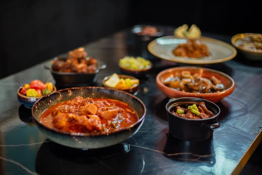 A black pan of red stew with chicken in the foreground, small bowls behind