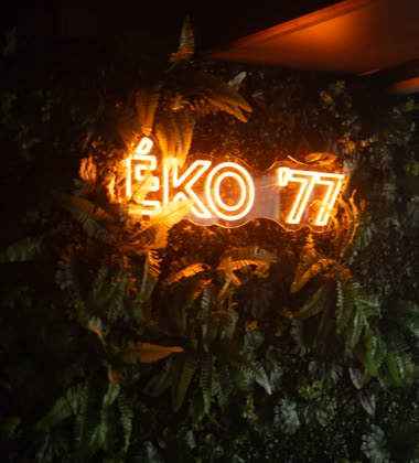 A neon ÉKO '77 sign glowing on a wall of ferns
