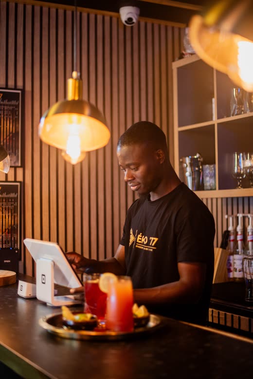 A member of staff at the bar in an ÉKO '77 T-shirt, a tray of iced drinks in front of him under a brass pendant lamp