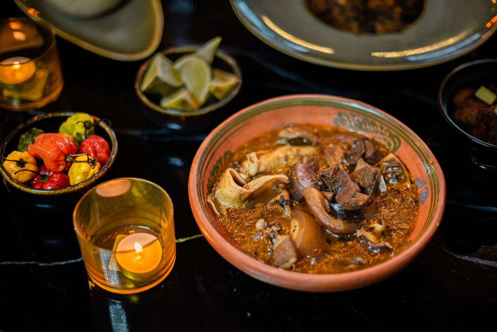 A bowl of pepper soup with assorted meat, with lime wedges and scotch bonnets alongside